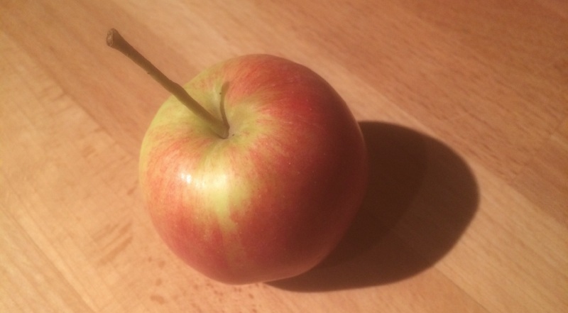 An red and green apple with a long stalk, casting a shadow onto the wooden suface it rests on.